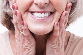 Nose-to-shoulder view of an older lady with mauve nails holding her cheeks and smiling